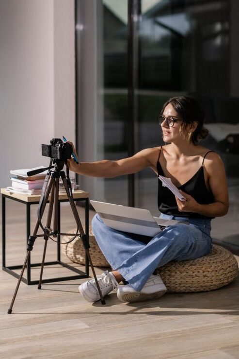 pexels photo 9040614 9040614 Woman sitting indoors with camera and laptop, engaging in vlogging activity.