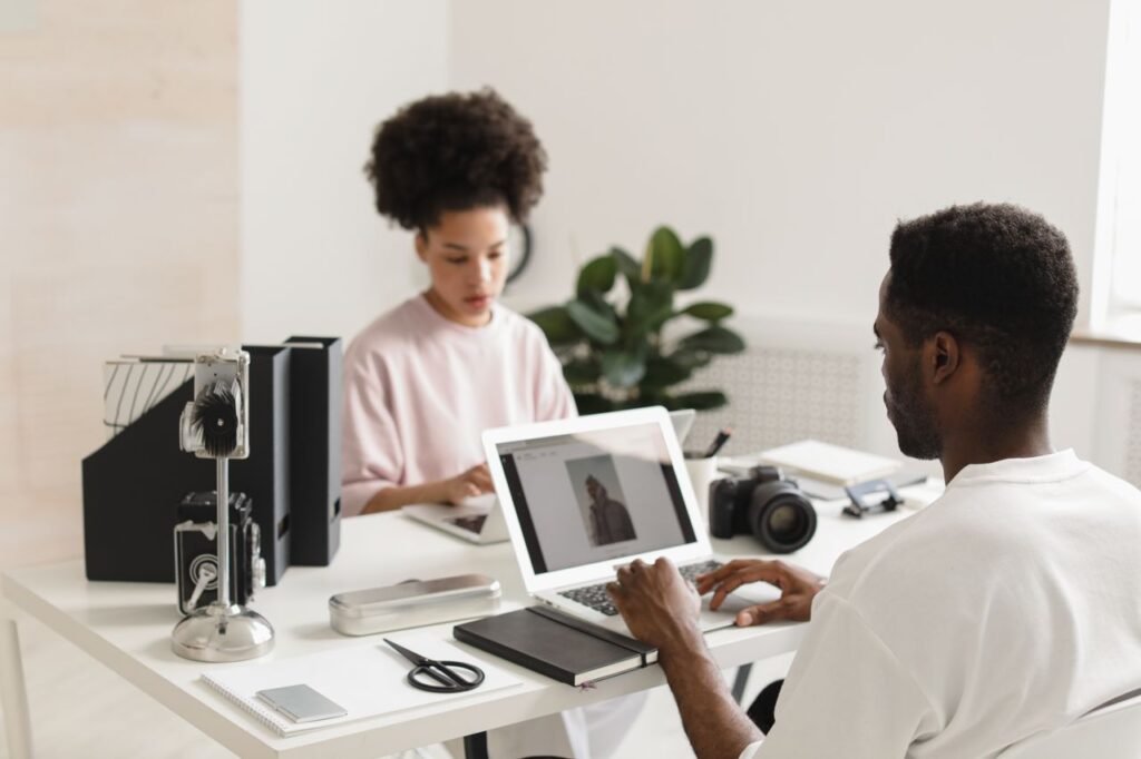 pexels photo 9909069 9909069 Two creative professionals collaborating in a bright, modern office setting with laptops and camera equipment.